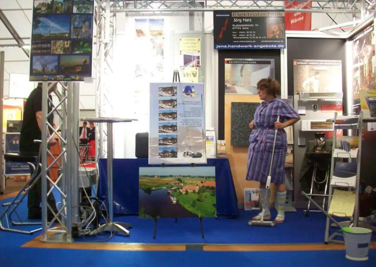 Moments Veranstaltungsagentur - Eine Frau in einem blau karierten Kleid posiert mit einem Wischmopp an einem Messestand in einer Halle.